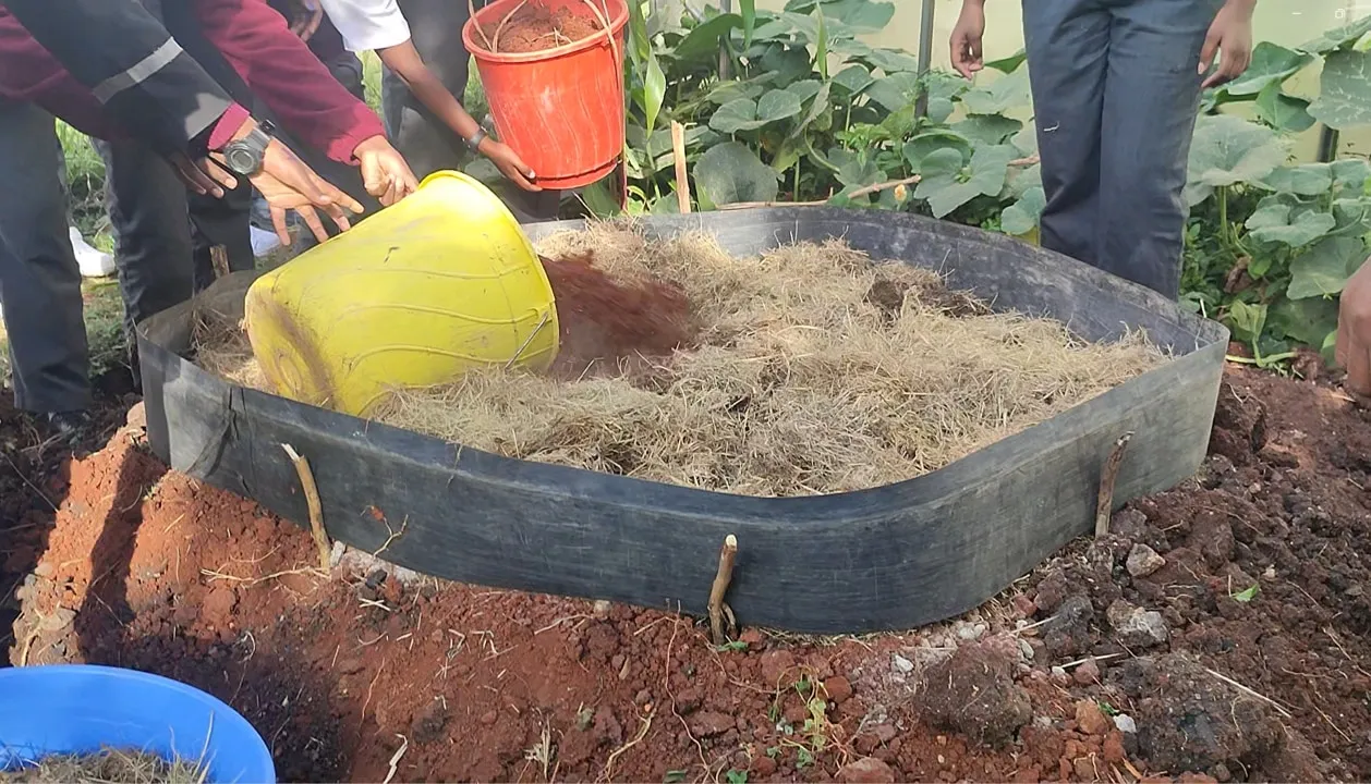 Students' hands layering dry grass and soil into a compost ring.